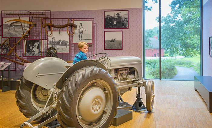 an old grey tractor is inside a museum gallery. A young boy with blond hair and a blue shirt sits on the tractor, as if driving it. There is a view ou